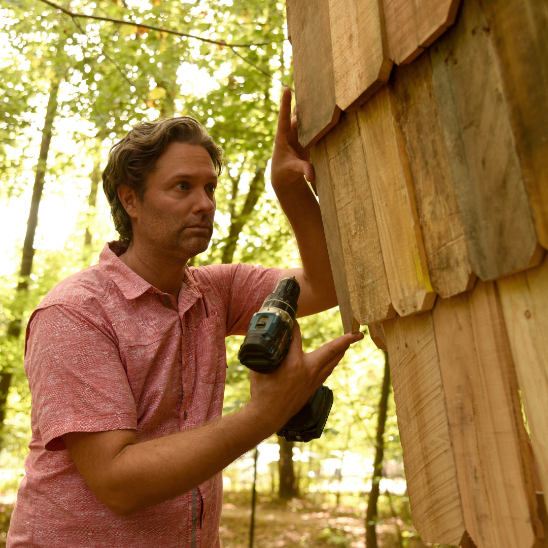 A whote man in a pink shirt holds a drill in his right hand as he positions a plank of wood on the side of a sculpture in the woods. 